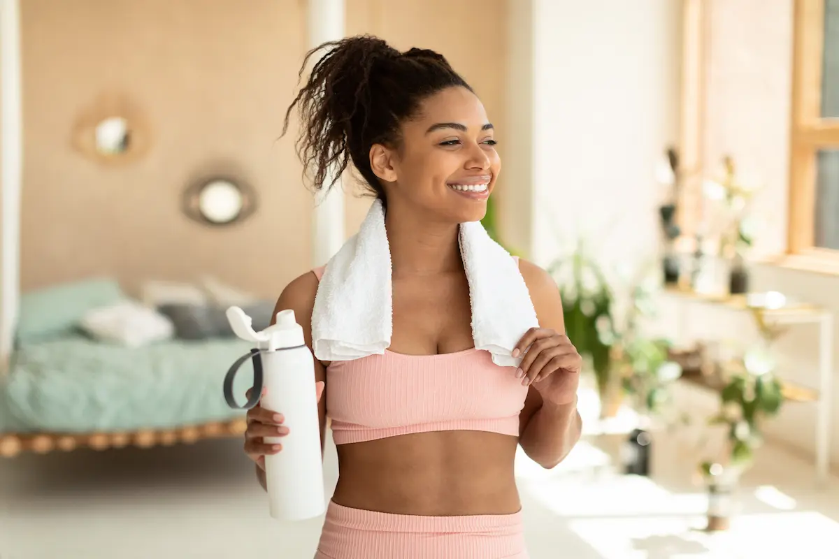 Mulher com roupa esportiva, toalha nos ombros e garrafa na mão, em imagem sobre liberdade e conforto com lentes de contato.